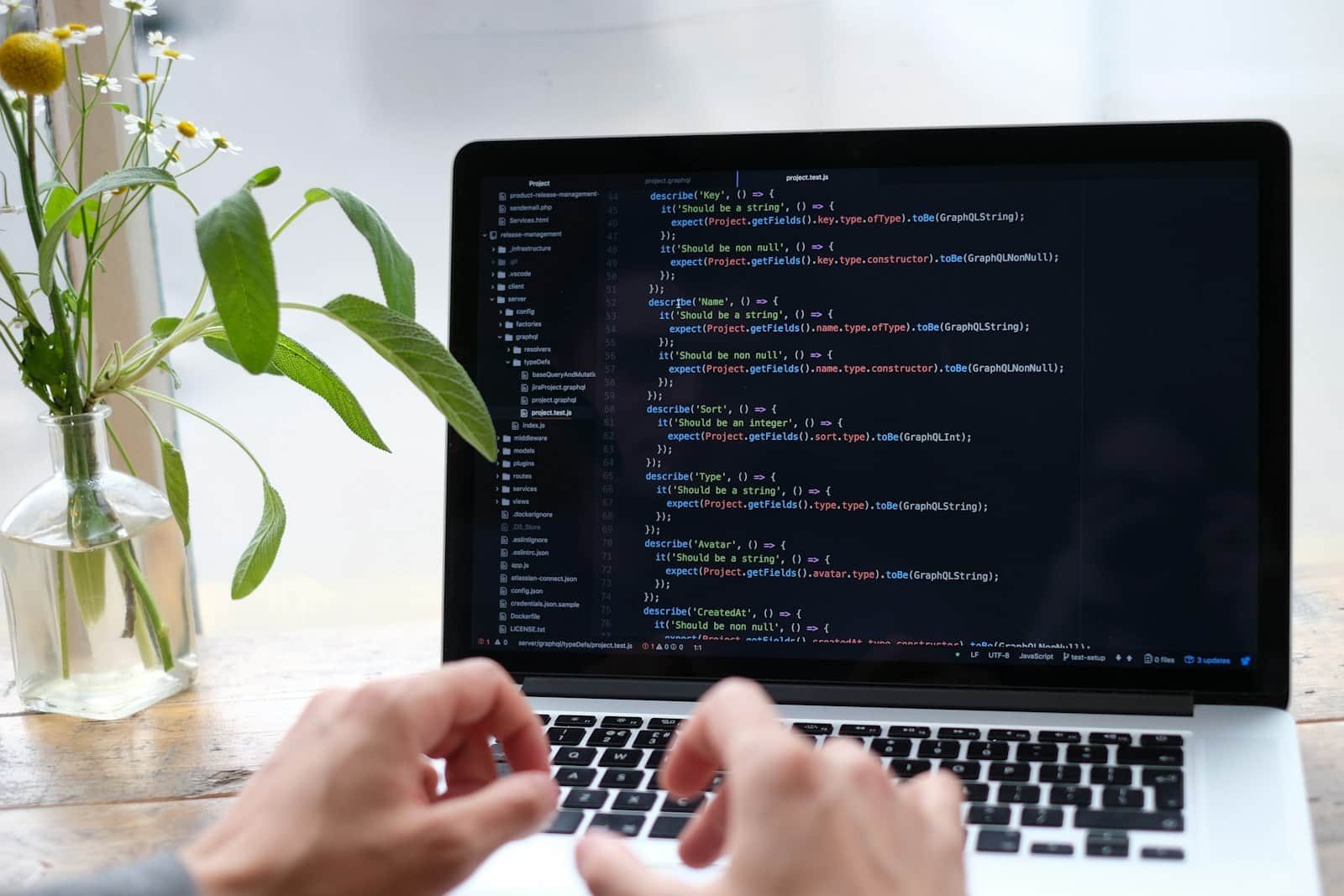 Hands writing code on a laptop in a bright workspace, flowers beside it